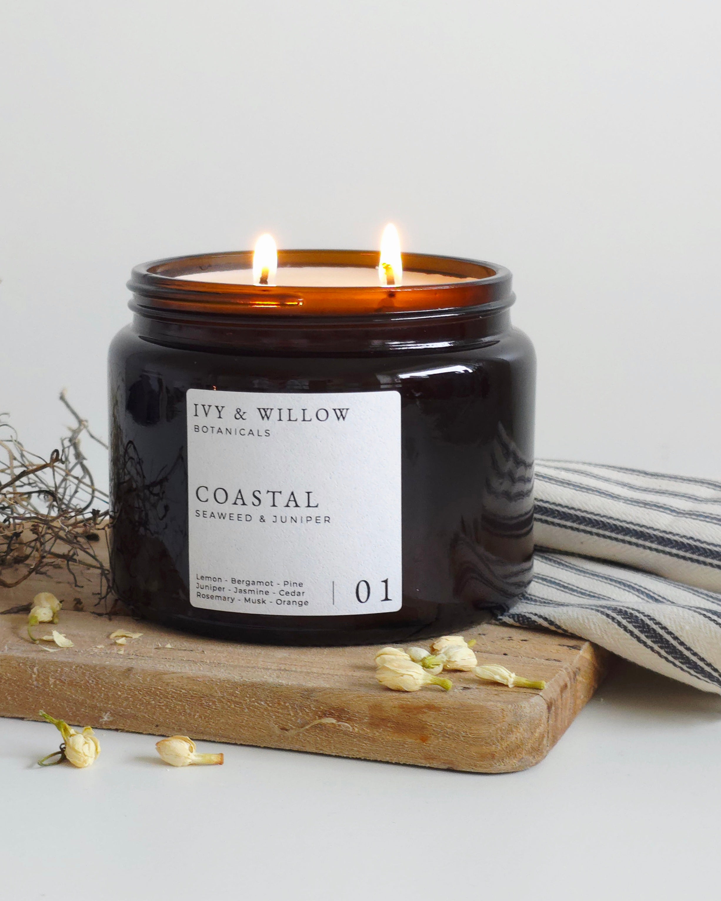 Black candle in a jar labeled 'Coastal' by Ivy & Willow Botanicals on a wooden board with dried flowers and a striped towel.