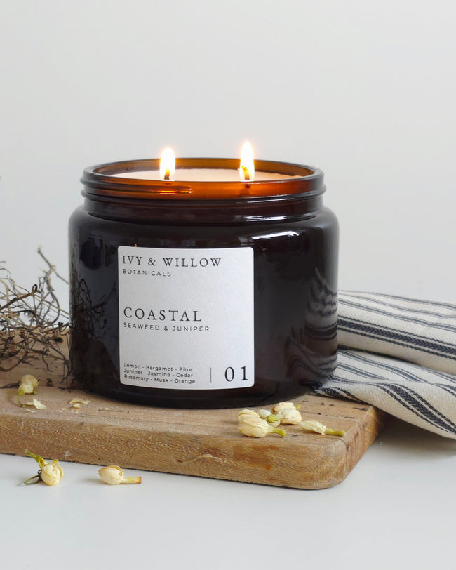 Black candle in a jar labeled 'Coastal' by Ivy & Willow Botanicals on a wooden board with dried flowers and a striped towel.