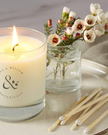 Candle with 'Fox & Willow Botanicals' branding, a vase of flowers, and matches on a surface.