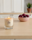 Candle in a glass jar on a kitchen counter with a bowl of plums and a plant in the background.