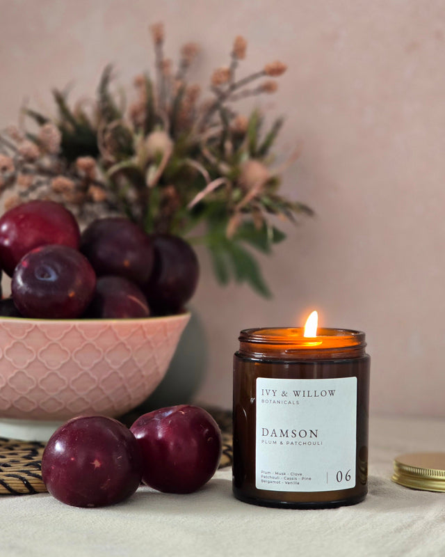 Lit candle with a label next to plums and a decorative bowl on a neutral background