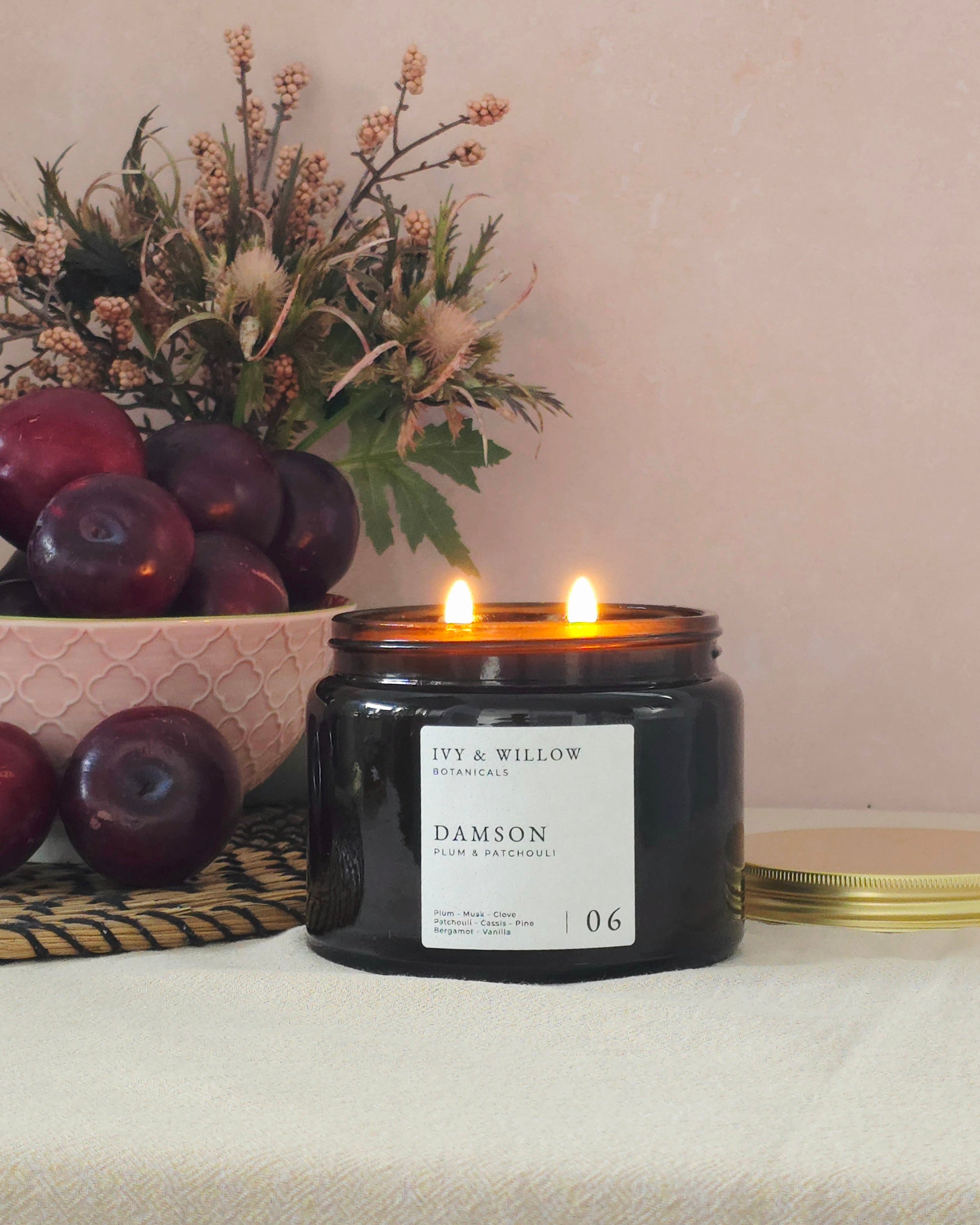 Candle with 'DAMSON' label on a table with apples and flowers.