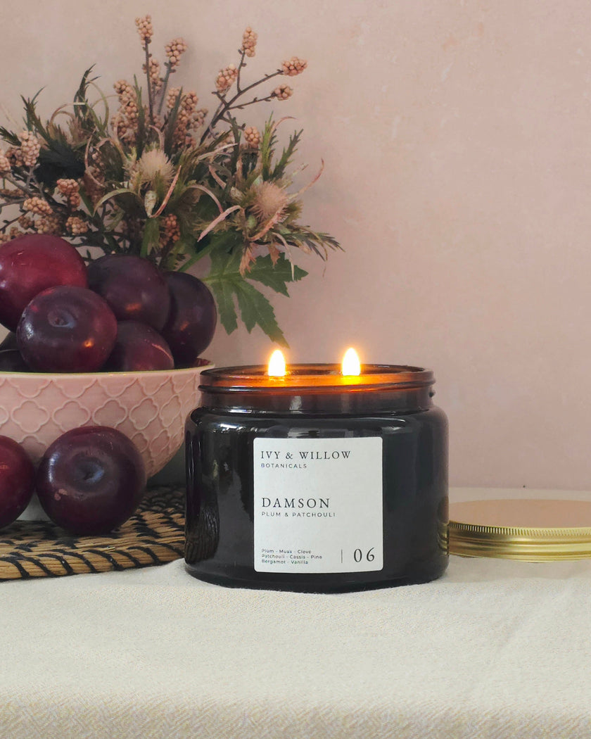 Candle with 'DAMSON' label on a table with apples and flowers.