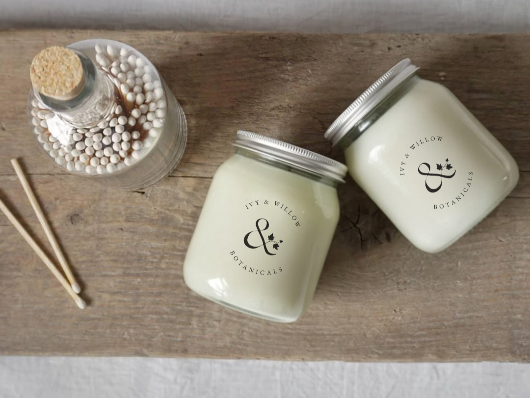 Two Signature candles from Ivy & Willow Botanicals on a wooden surface with a small bottle and sticks.