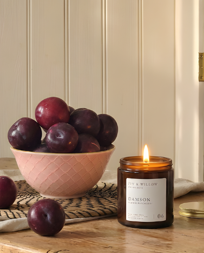 Bowl of plums next to a lit candle labeled 'Damson' on a wooden surface.