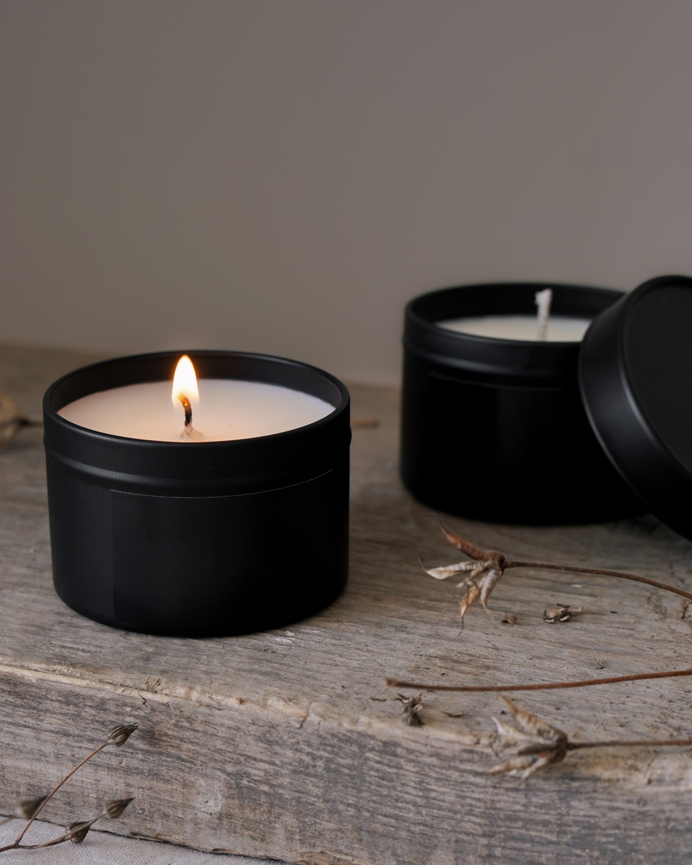 Two black candles in tins on a wooden surface with a neutral background
