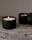 Two black candles in tins on a wooden surface with a neutral background