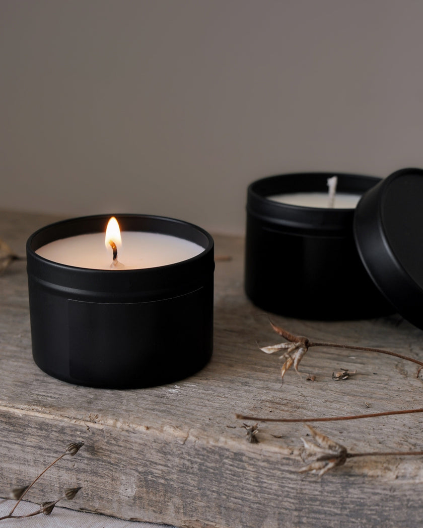 Two black candles in tins on a wooden surface with a neutral background
