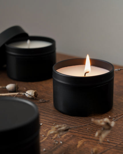 Black candle in a tin with a lit wick on a wooden surface, surrounded by dried plants.