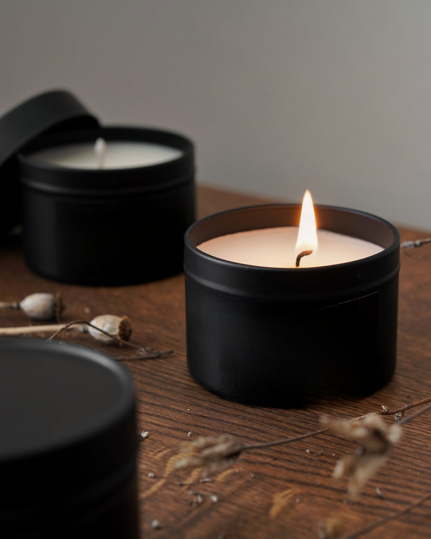 Black candle in a tin with a lit wick on a wooden surface, surrounded by dried plants.