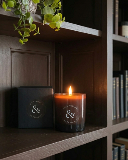 Candle with a logo on a shelf next to a black box, with a plant and books in the background.