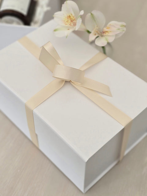 White gift box with a beige ribbon and flowers on a light surface