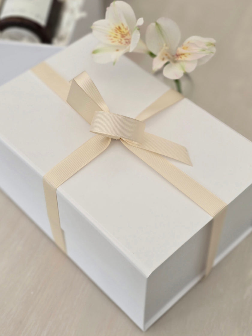 White gift box with a beige ribbon and flowers on a light surface