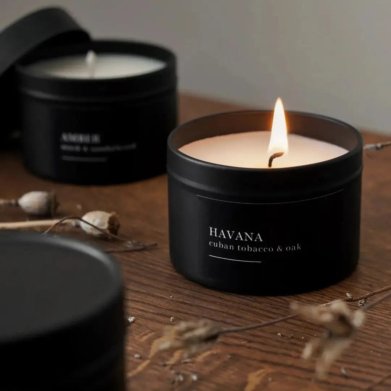 Candle in a black container labeled 'Havana' on a wooden surface with another candle in the background.
