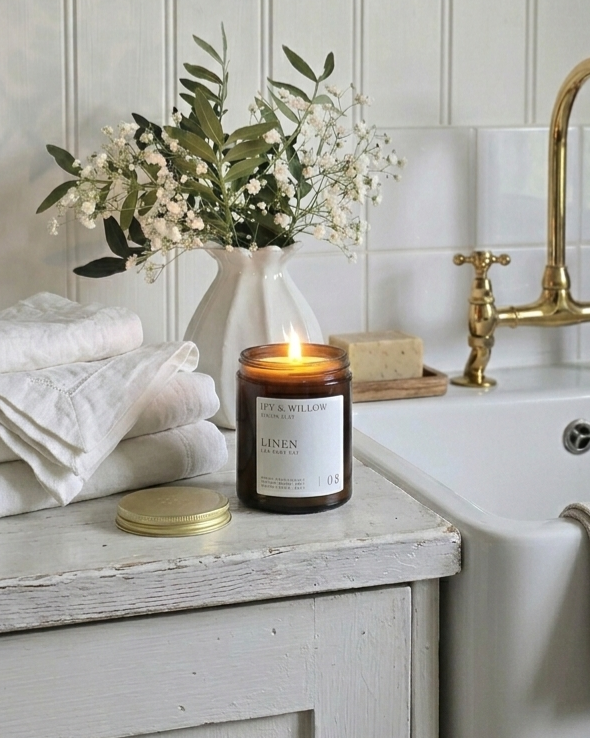 Candle with a label on a wooden surface next to a vase of flowers and a sink.