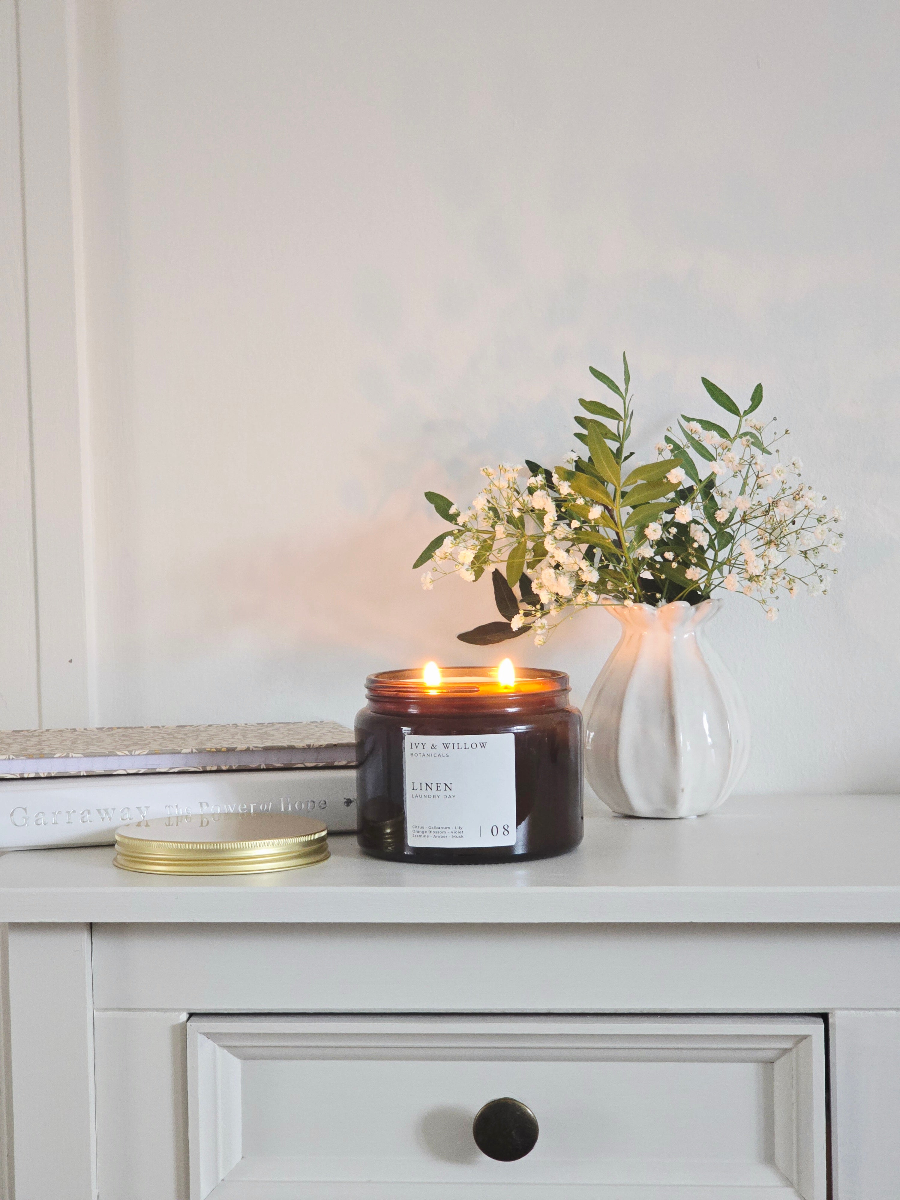 Candle with a label on a white surface next to a vase with flowers