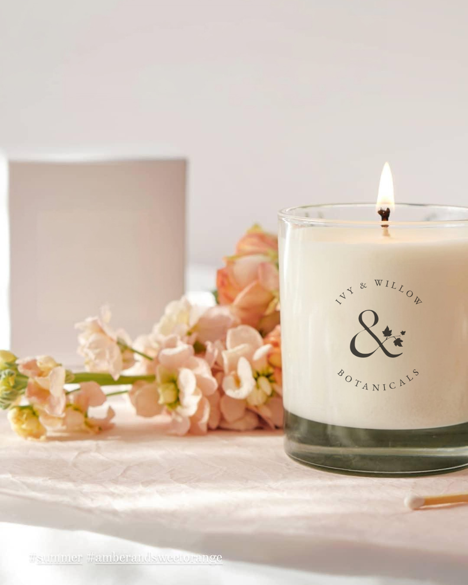 Candle with 'Ivy & Willow Botanicals' logo next to flowers on a white surface