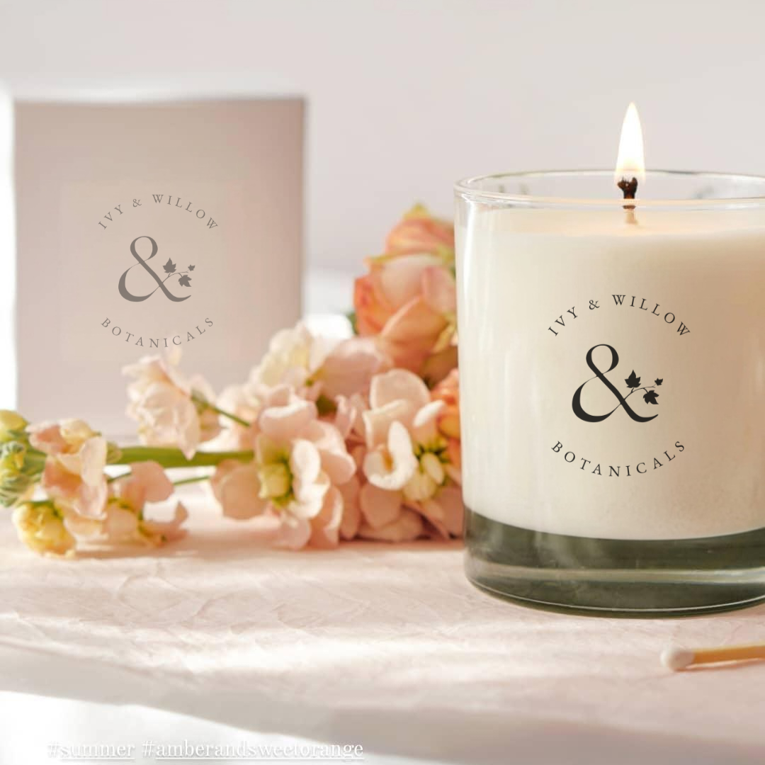 Candle with 'Ivy & Willow Botanicals' branding on a surface with flowers and a box in the background.