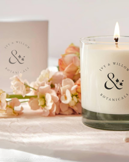 Candle with 'Ivy & Willow Botanicals' branding on a surface with flowers and a box in the background.