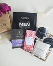 Collection of men's skincare products and tea packages on a white surface with a floral touch.