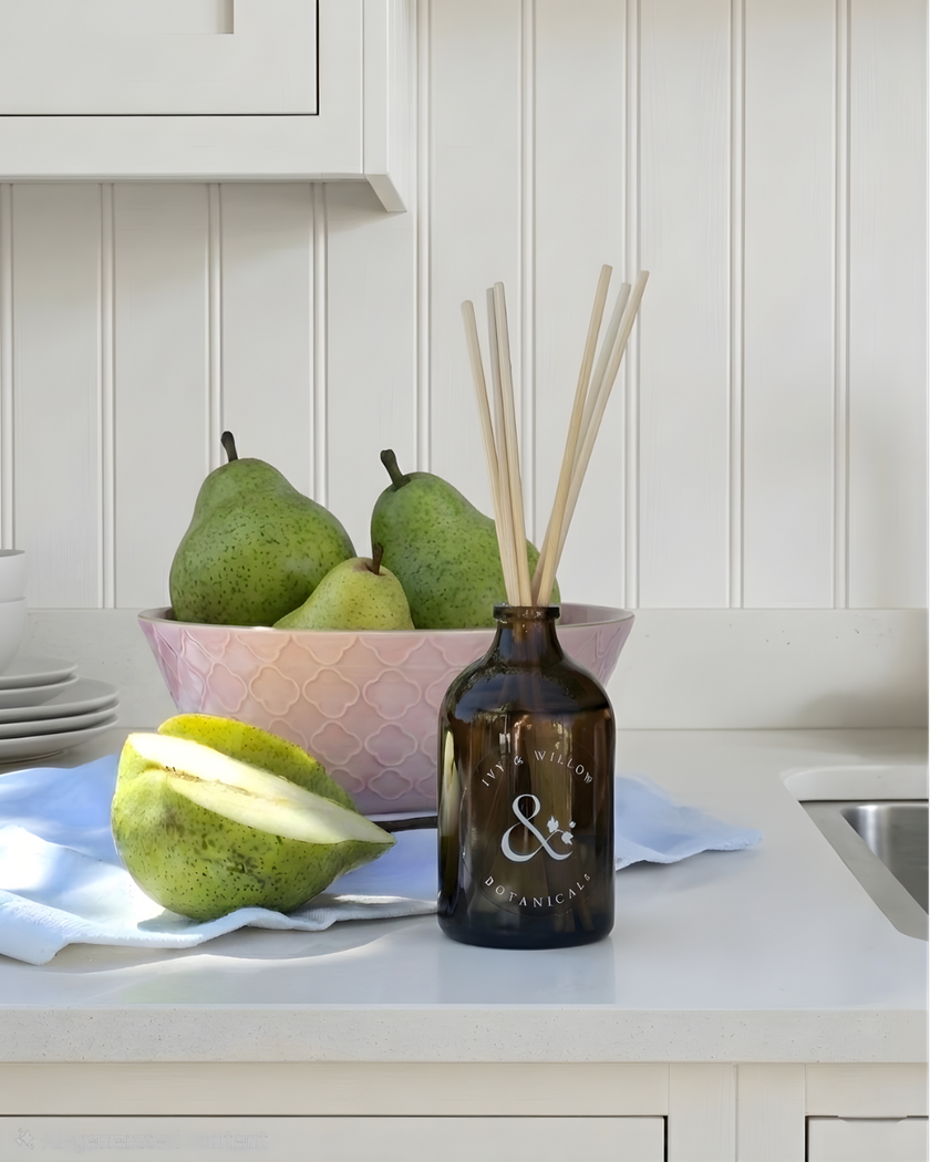 Bottle of diffuser with reeds on a kitchen counter with pears and a bowl.