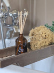 Brown diffuser bottle with wooden sticks on a wooden tray in a bathroom setting.