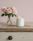Candle labeled 'Spring' with a vase of pink roses on a wooden surface.