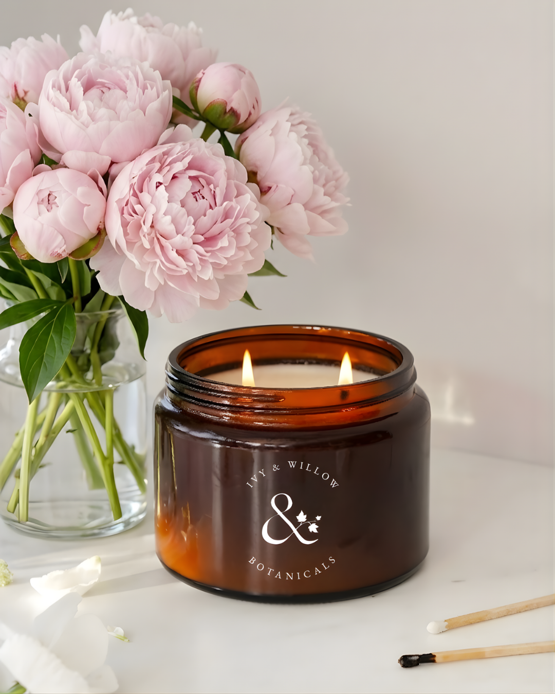 Candle in an amber jar with a floral peony arrangement on a light surface