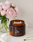 Candle in an amber jar with a floral peony arrangement on a light surface