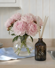 Pink flowers in a clear vase on a kitchen counter with a dark brown bottle labeled 'and.'