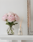 Vase of pink flowers and diffuser on a white surface with a decorative frame in the background