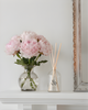 Vase of pink flowers and diffuser on a white surface with a decorative frame in the background