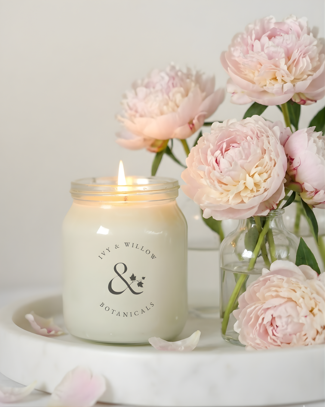 Lit candle in a jar with 'Ivy & Willow Botanicals' branding next to pink Peony flowers.