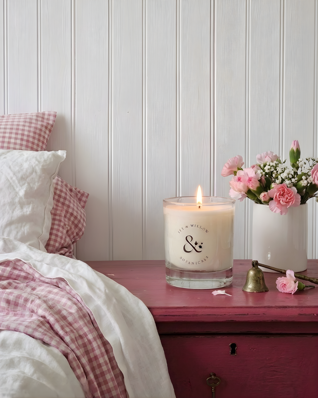 Candle with a logo on a wooden surface next to a bed with pink and white checkered pillows.