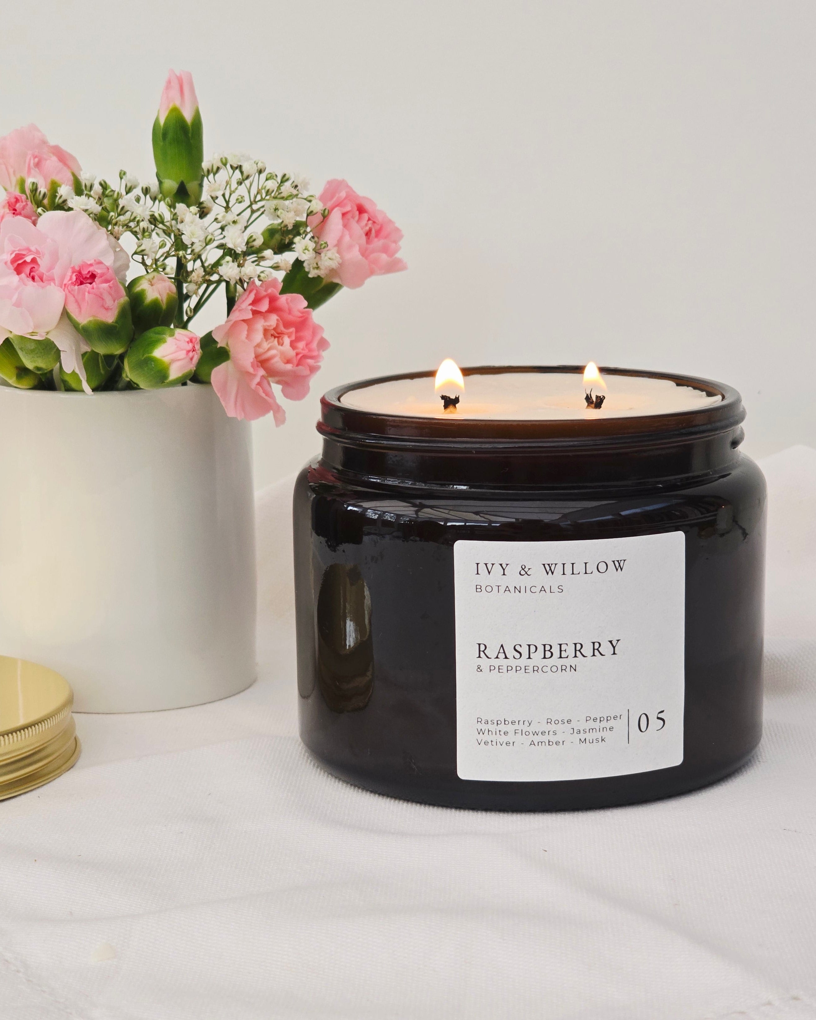 Black candle jar with a label on a white surface next to pink flowers