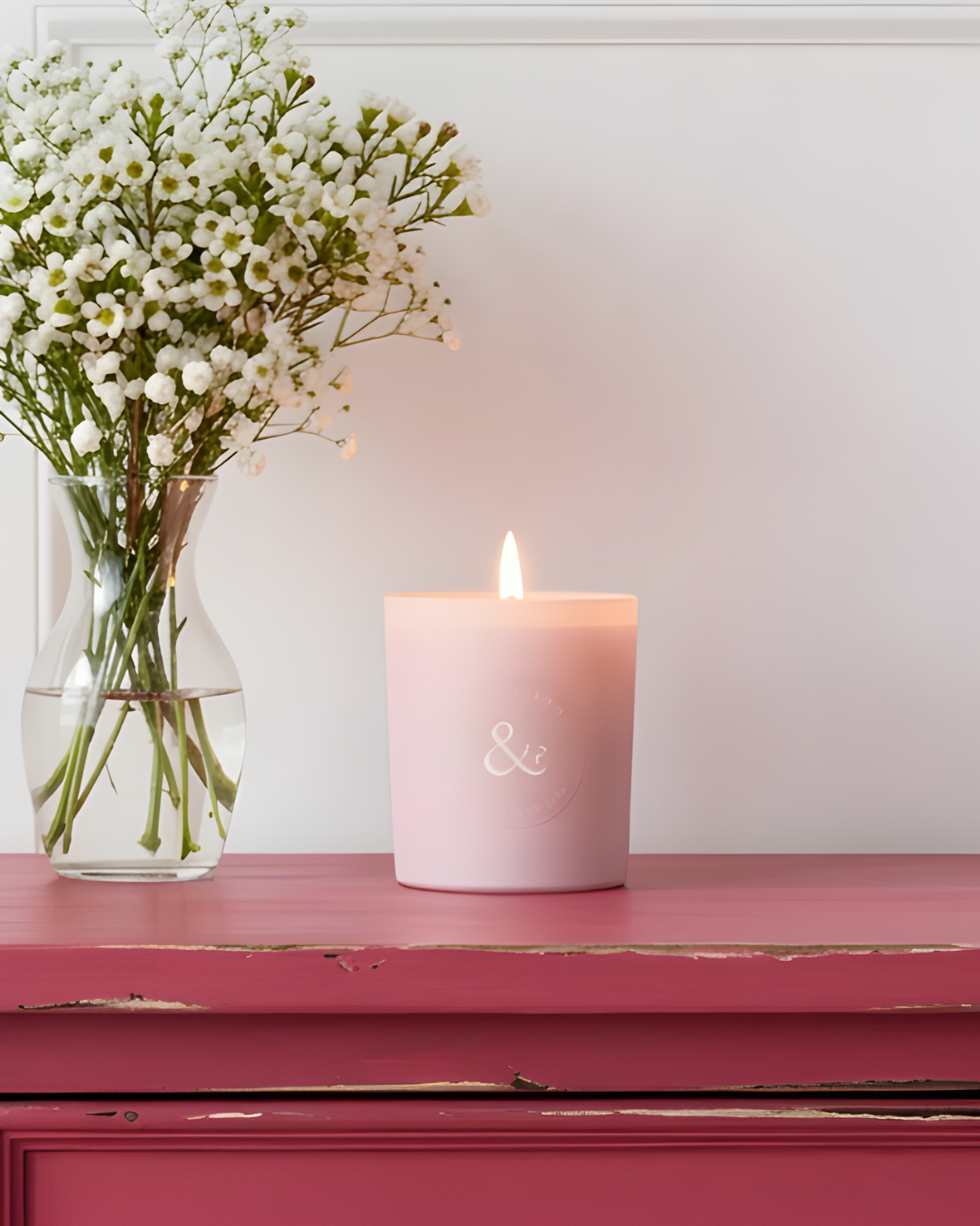 Pink candle with a small flame next to a vase of white flowers on a red surface.