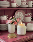 Candle on a pink surface with floral arrangement and patterned dishes in the background