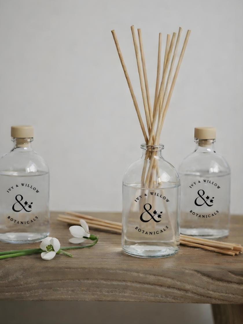 Two clear bottles with bamboo sticks and flowers on a wooden surface, featuring 'ivy & willow botanics' branding.