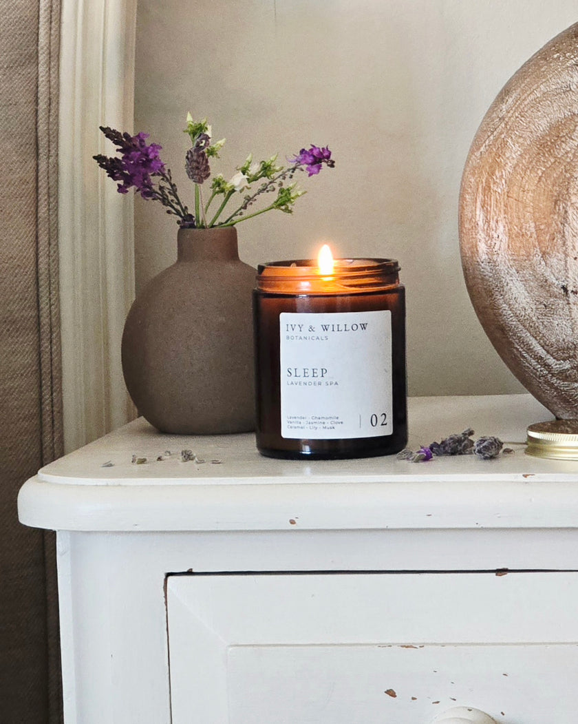 Candle labeled 'Sleep' by 'Lily & Willow' on a white surface with a vase of flowers and a decorative sphere.