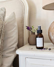 Bottle of room spray on a nightstand with a vase of flowers and lamp in the background.