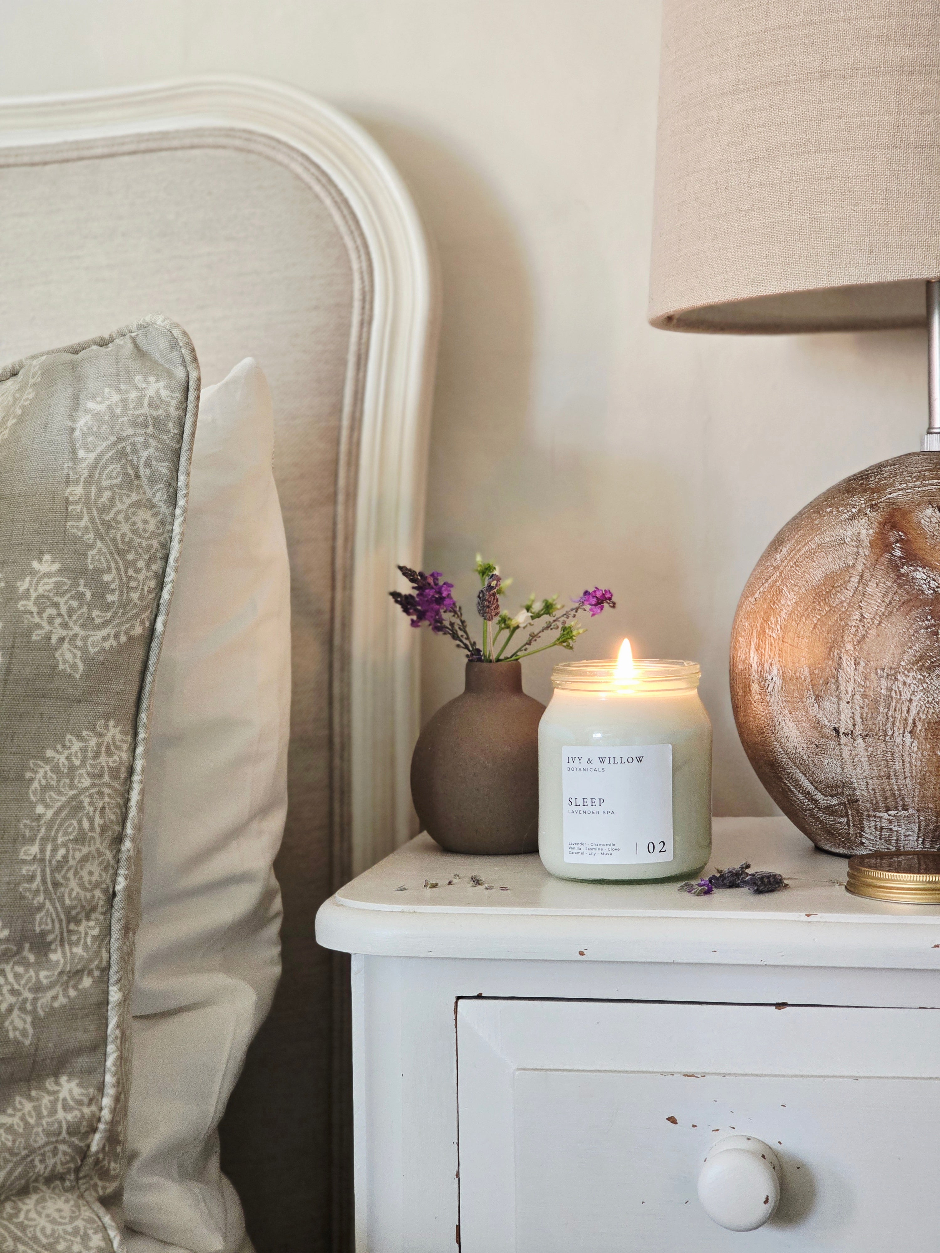 Candle on a nightstand next to a lamp and decorative vase with flowers.