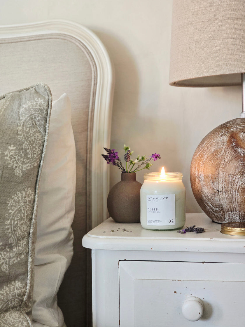 Candle on a nightstand next to a lamp and decorative vase with flowers.