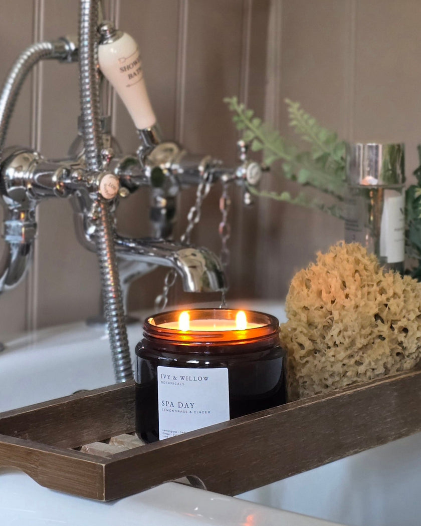 Candle in a black container on a wooden tray with a bathtub and faucet in the background