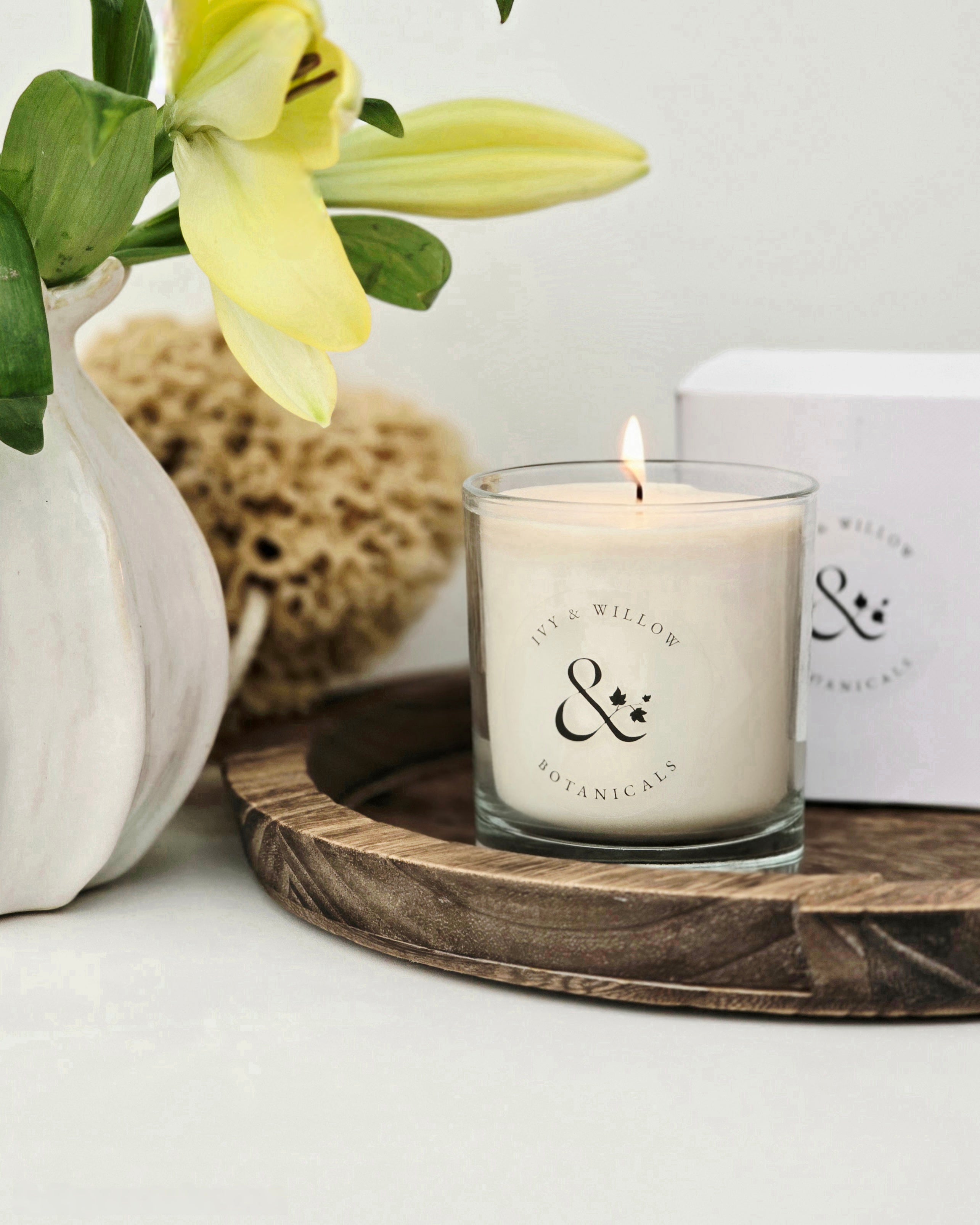 Candle with a logo on a wooden tray with flowers and a box in the background