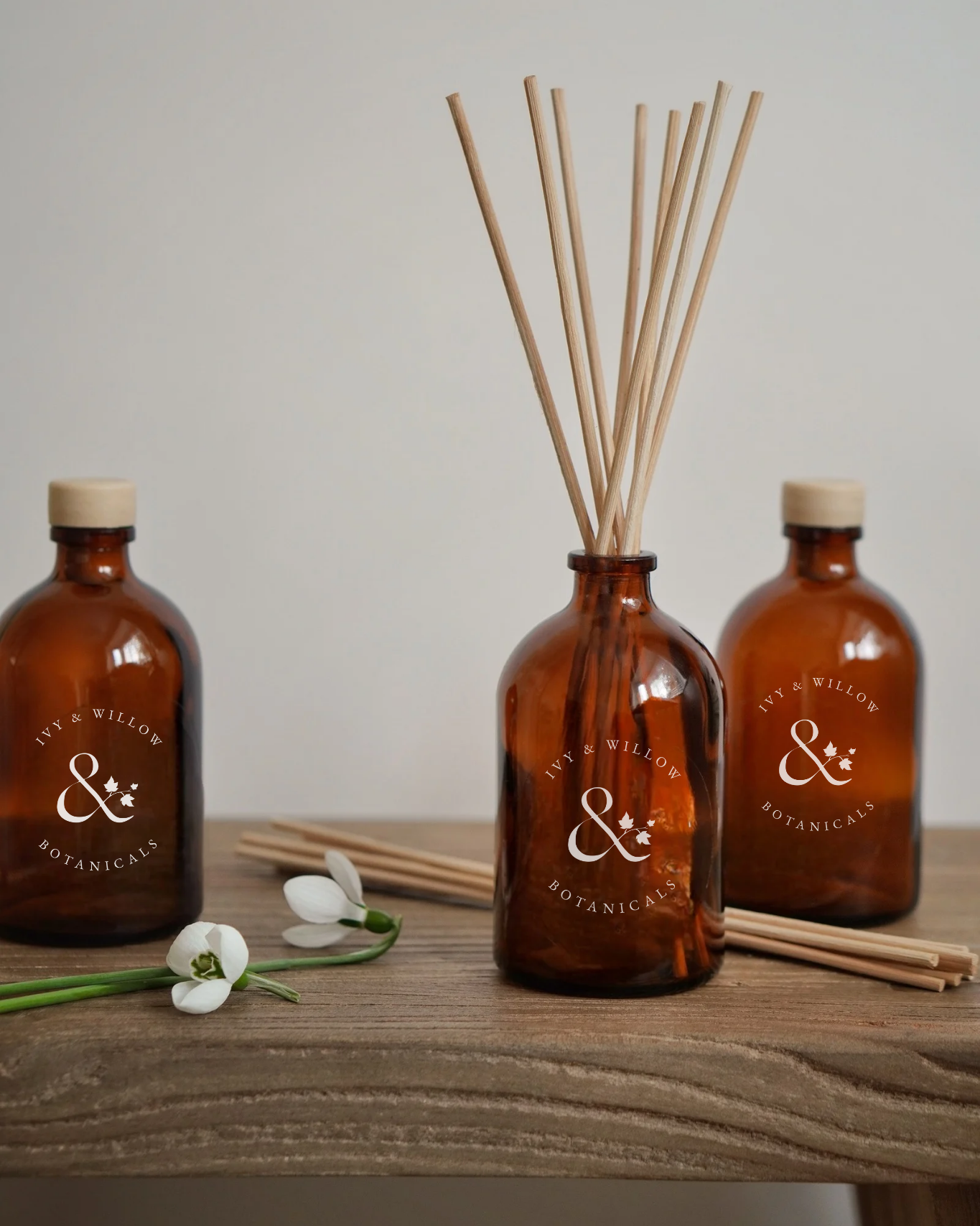 Three amber glass bottles with reeds and a white flower on a wooden surface.
