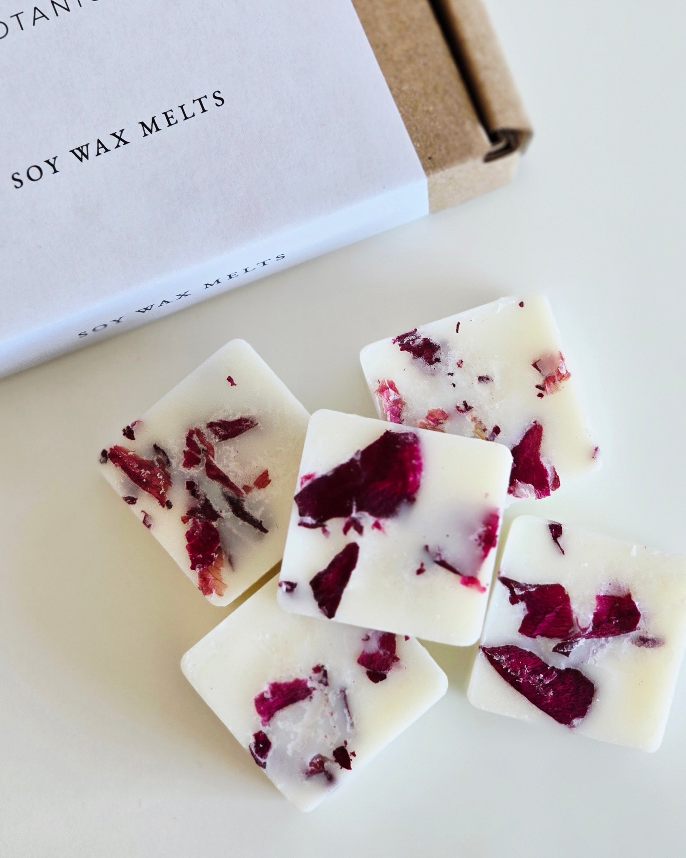 Soy wax melts with red flower petals on a white surface, next to a box labeled 'Soy Wax Melts'.