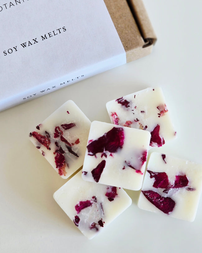 Soy wax melts with red flower petals on a white surface, next to a box labeled 'Soy Wax Melts'.