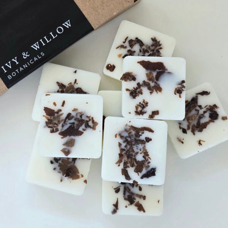 White soap bars with dark specks on a white surface, with 'Ivy & Willow Botanicals' packaging in the background.