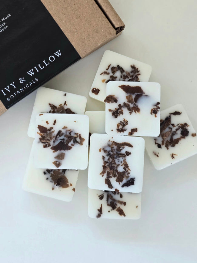 White soap bars with dark specks on a white surface, with 'Ivy & Willow Botanicals' packaging in the background.