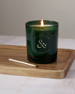 Green candle with a lit wick on a wooden tray against a neutral background